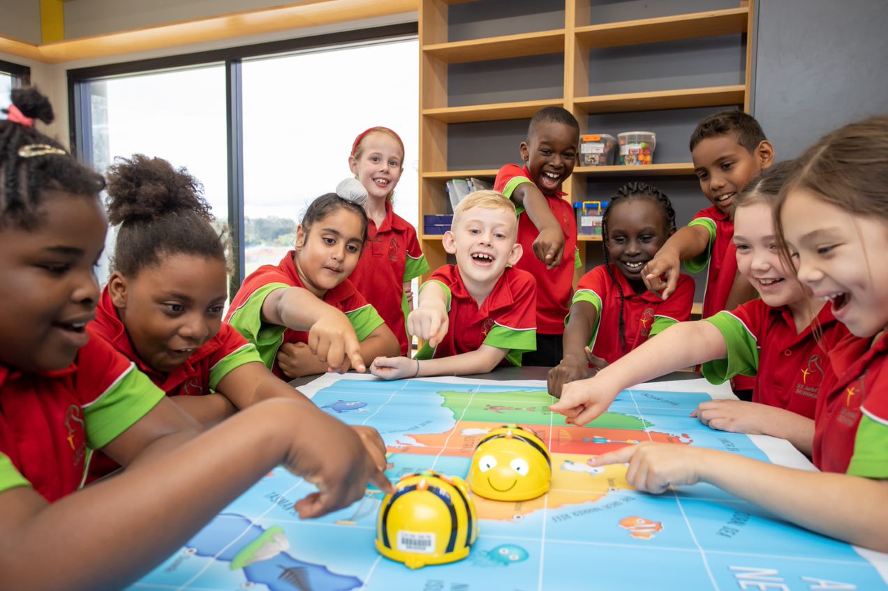 Students gathered around a table pointing at a map while using small programmable robots during a classroom activity