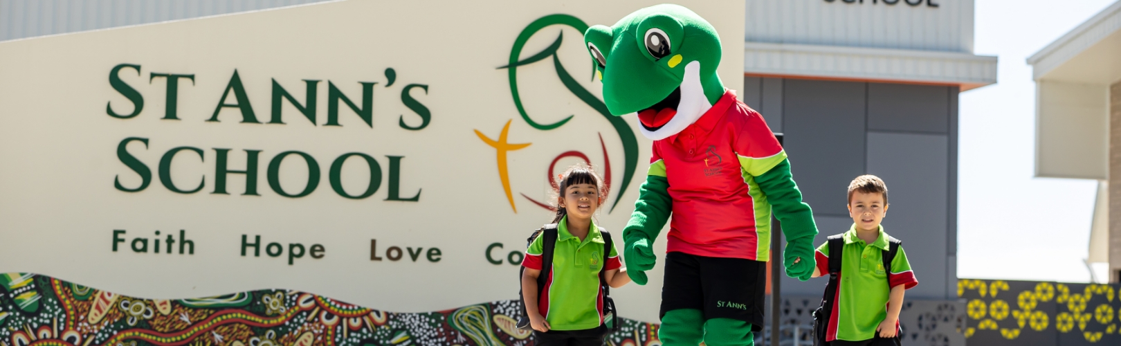 School mascot in a green costume walking with two students in front of a “St Ann’s School” sign with the words “Faith Hope Love” visible